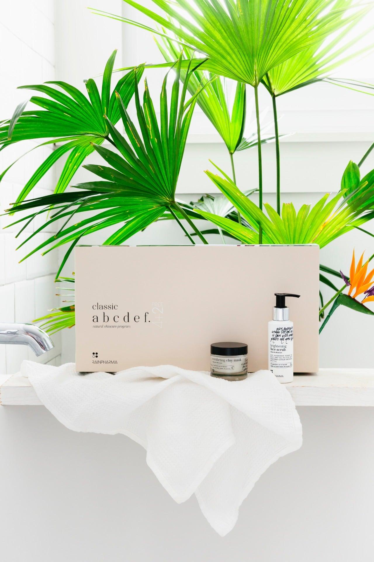 De Complete Huidverzorgingsset - abcdef promobox 4+2 GRATIS van RainPharma en een witte handdoek worden uitgestald op een plank voor een plantenachtergrond, die een holistische benadering tot natuurlijke huidverzorging omarmt.