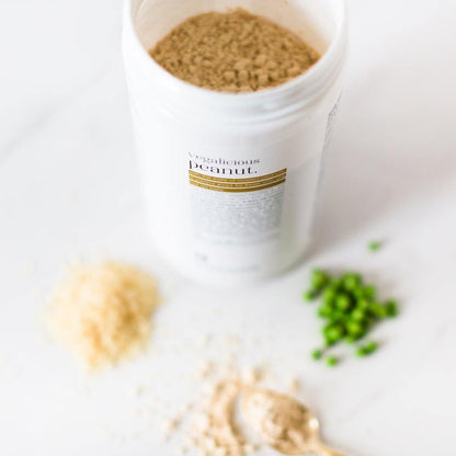 Een potje Vegalicious Peanut Shake van Rainpharma met het opschrift &quot;Vegalicious Peanut Shake&quot; op een witte ondergrond, met een lepel poeder en gehakte groene erwten ernaast, is een gluten- en caseïnevrij genot.