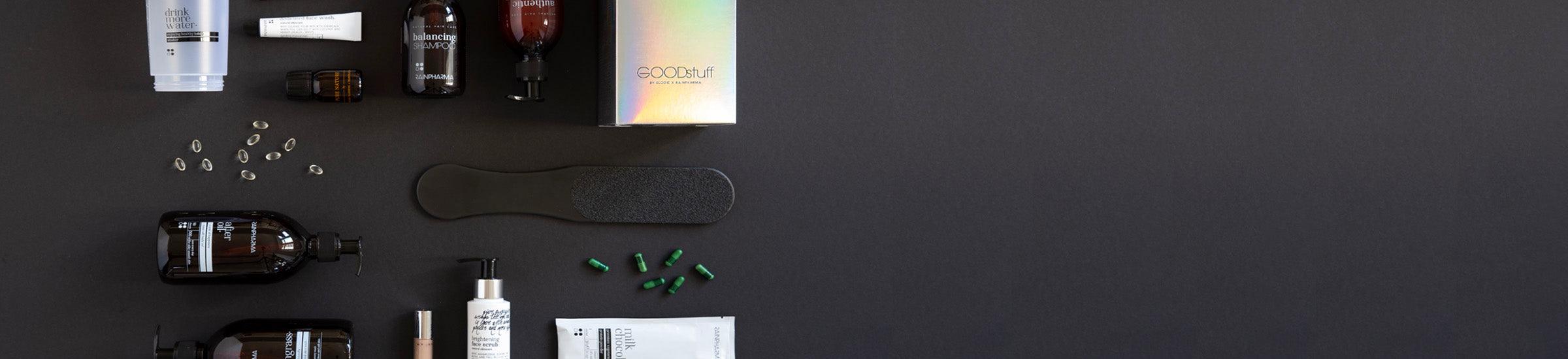 Verschillende huidverzorgings- en verzorgingsproducten liggen op een zwart oppervlak, waaronder flesjes, een puimsteen en kleine capsules.