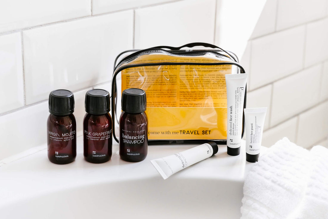 Een RainPharma Travelset Come With Me, uitgestald op de rand van een badkuip, bevat producten in reisformaat: kleine flesjes shampoo, mondwater, grapefruitwasmiddel, tubes lotion en crème, en een doorzichtige en gele draagtas.