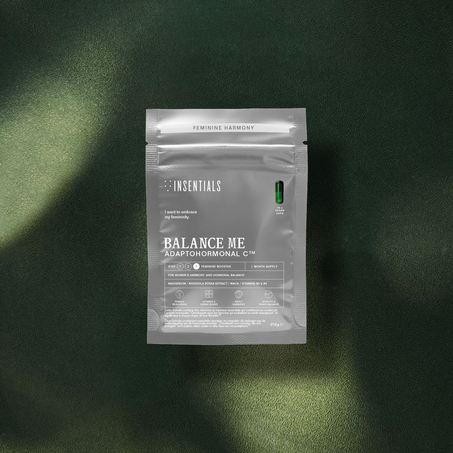Een zilverkleurig zakje met het opschrift „Balance Me – Hormonale Balans &amp; Menopauze Support” van Insentials ligt op een donkergroen, gestructureerd oppervlak, met daarbovenop één groene capsule.