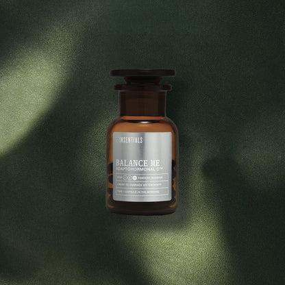 Een bruine glazen fles Insentials Balance Me – Hormonale Balans &amp; Menopauze Support met maca en rhodiola staat op een groen oppervlak met structuur, badend in gevlekt licht, wat de indruk wekt van natuurlijke hormonale regulatie.