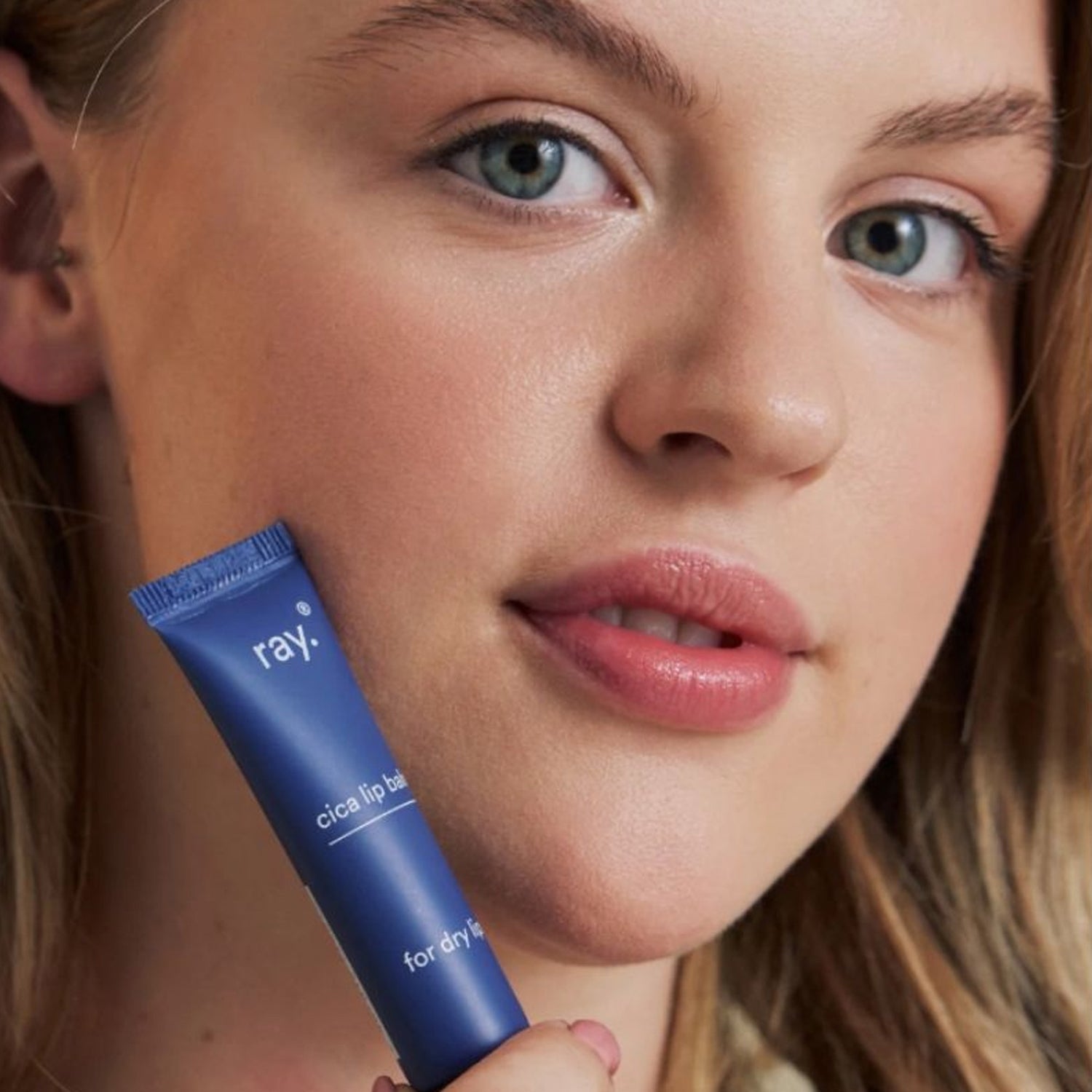 Iemand met een lichte huid houdt een blauwe tube Ray Cica Lip Balm bij de lippen - ideaal voor verzachtende verzorging van droge lippen met madecassoside.