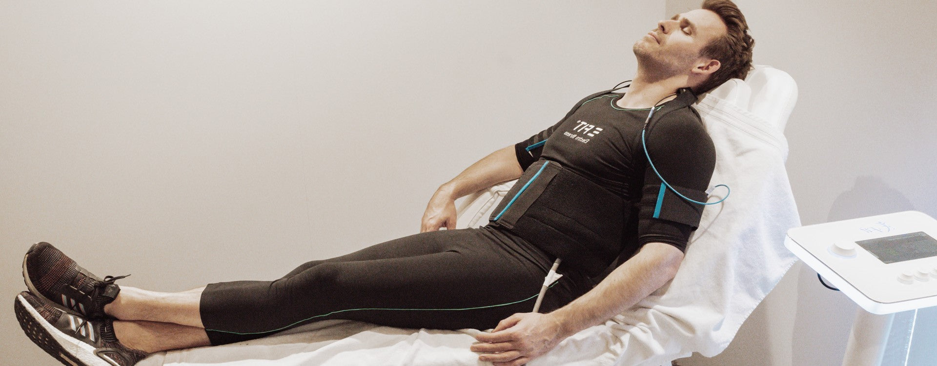 Een man in atletische kleding ligt op een ligstoel met elektroden en draden aan zijn lichaam bevestigd en ondergaat een behandeling met elektrische spierstimulatie.
