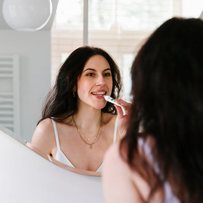 Een vrouw staat voor de spiegel en gebruikt een elektrische tandenborstel. Ze draagt een wit topje en meerdere kettingen in verschillende lagen. Haar glimlach wordt versterkt door de lippenbalsem van RainPharma, verrijkt met hydraterende plantaardige oliën.