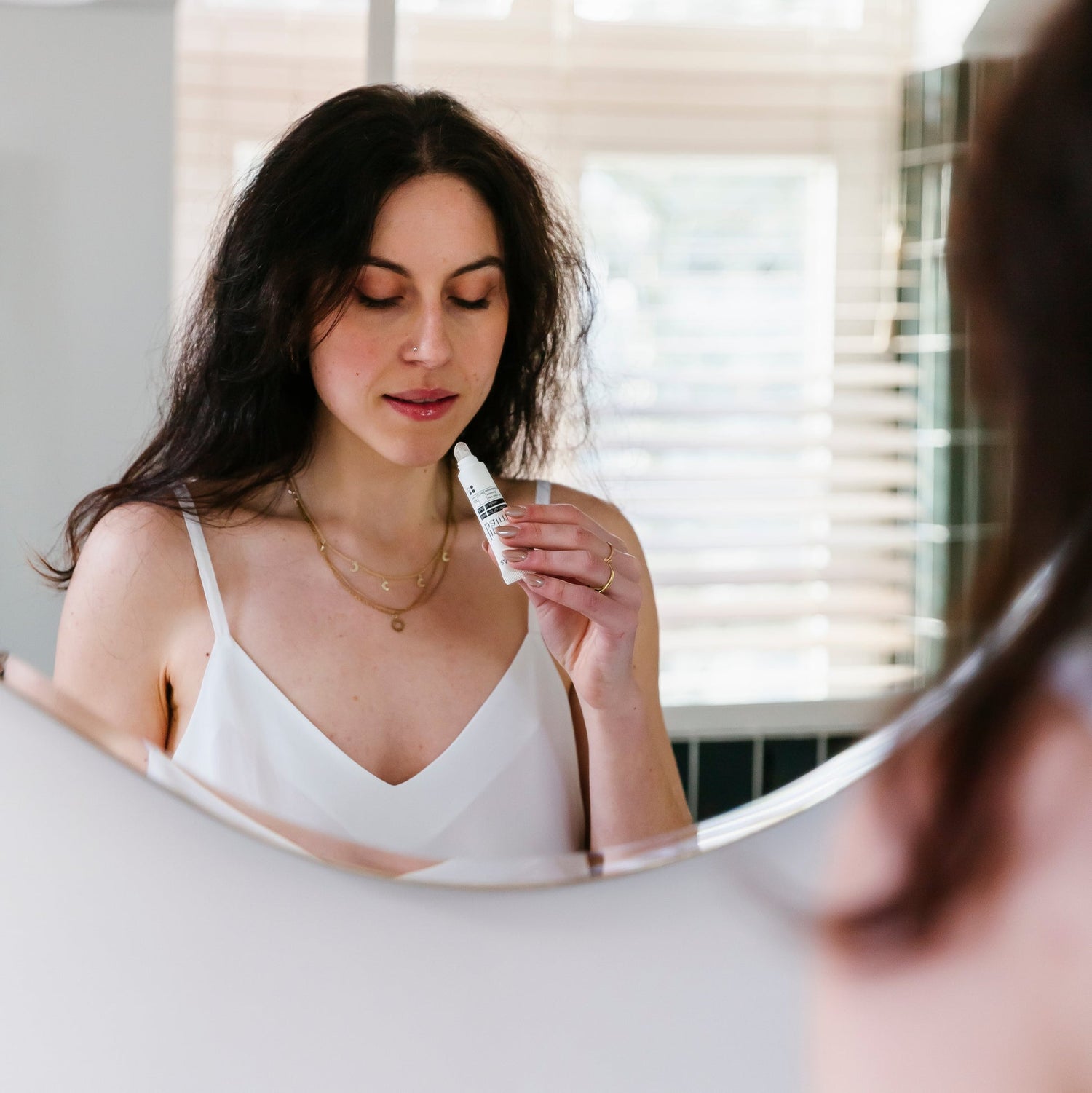 Een persoon staat voor een spiegel met RainPharma lippenbalsem, gekleed in een wit hemdje en meerdere kettingen.