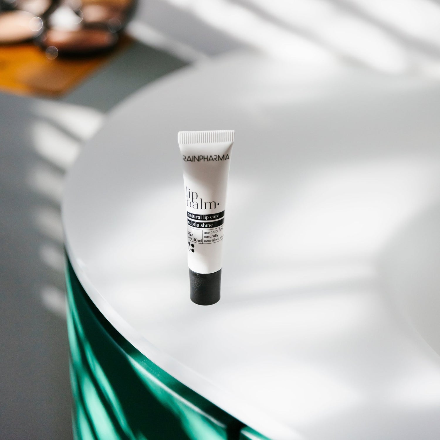 Een tube RainPharma Lippenbalsem ligt op een wit, rond oppervlak, terwijl zonlicht patronen creëert.