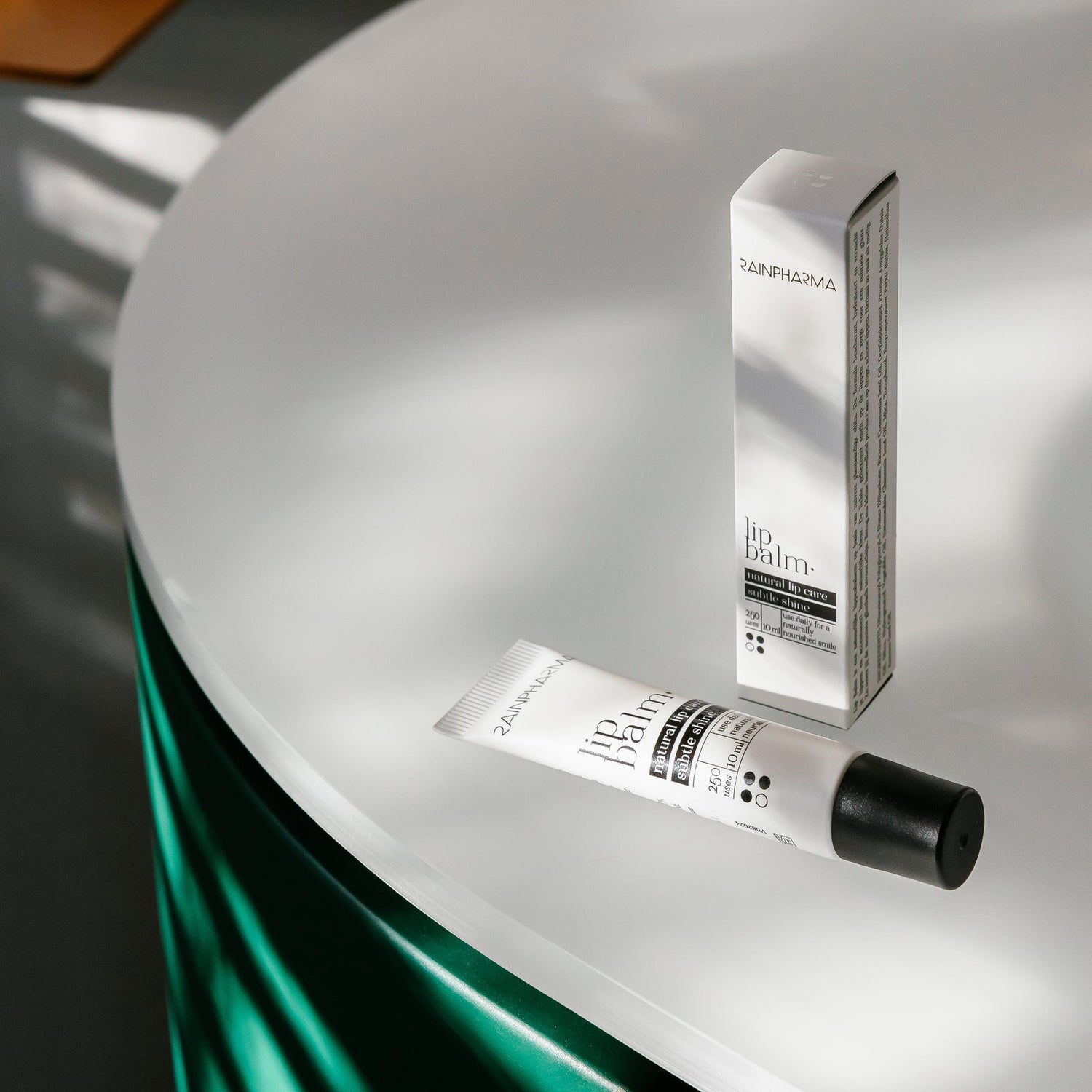 Een RainPharma lippenbalsem, verrijkt met hydraterende plantaardige oliën, is afgebeeld op een rond, wit oppervlak tegen een achtergrond van groene stof.