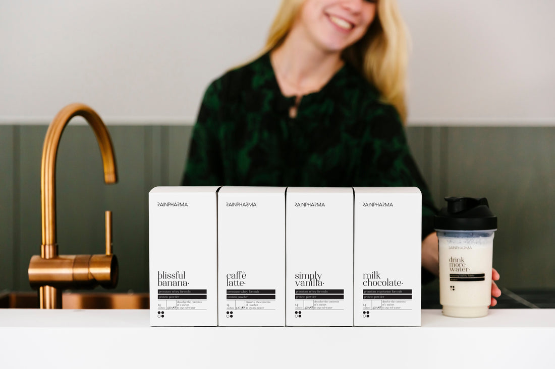 Vier witte dozen RainPharma Shake Blissful Banana Zakjes, een GMO-vrije proteïnedrankmix, staan op een witte toonbank opgesteld voor een vrouw met een shakerbeker naast een gouden kraan.