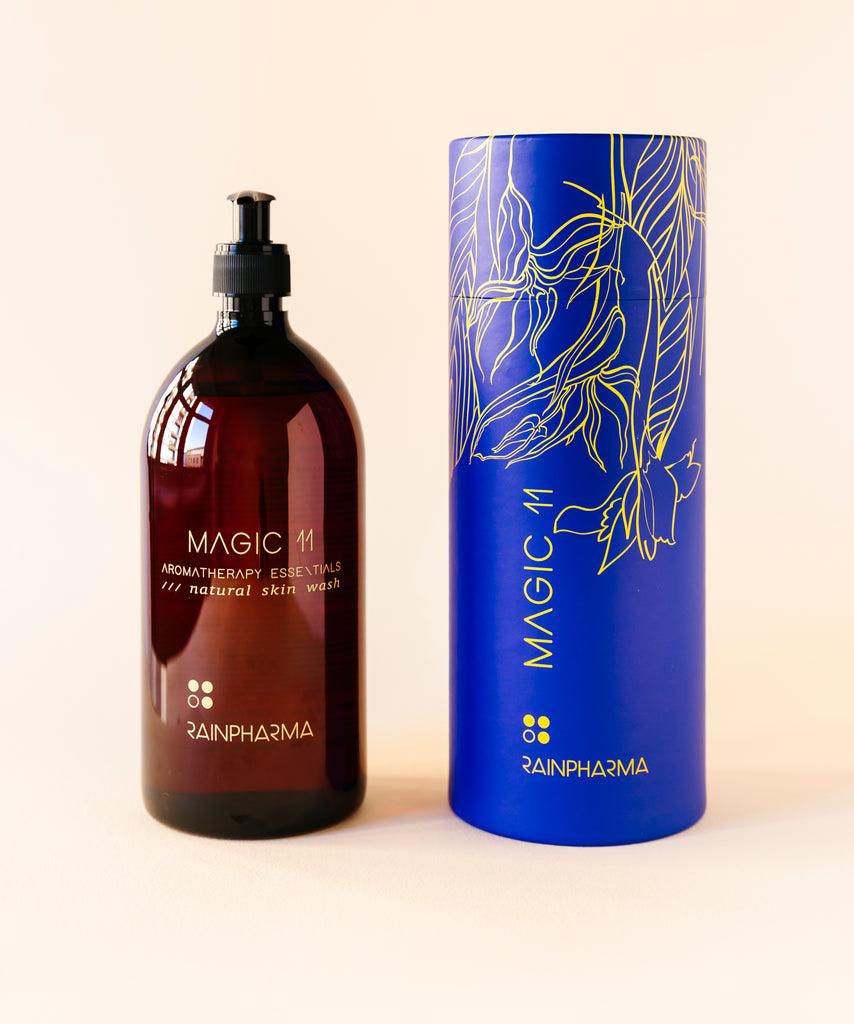 Een bruine fles Skin Wash Magic 11 van RainPharma, verrijkt met essentiële oliën, staat naast een blauwe cilindervormige verpakking met een gouden bloemmotief.