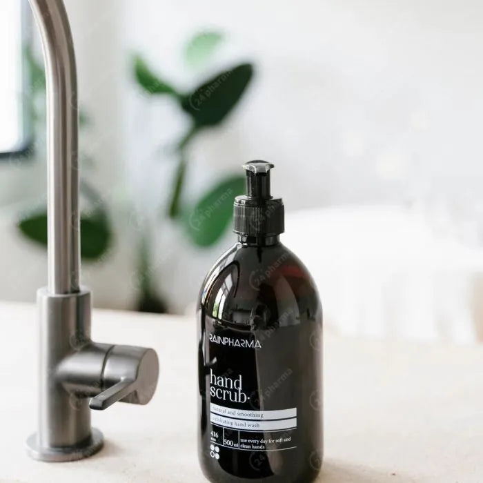 Een zwarte RainPharma Hand Scrub - 500 ml fles met bamboe scrubdeeltjes rust op een wit aanrecht naast een zilveren kraan, met een vage groene plant op de achtergrond.