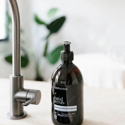 Een zwarte RainPharma Hand Scrub - 500 ml fles met bamboe scrubdeeltjes rust op een wit aanrecht naast een zilveren kraan, met een vage groene plant op de achtergrond.