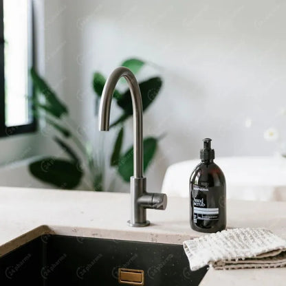 Een modern aanrecht met een gebogen metalen kraan, een 500 ml RainPharma Hand Scrub fles en een textieldoek op het aanrecht; op de achtergrond zijn planten te zien.
