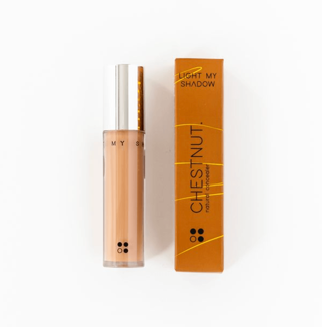 Een tube vloeibare concealer "Light my shadow" van RainPharma staat naast een feloranje doos met het label "Chestnut", wat de veganistische formule benadrukt.