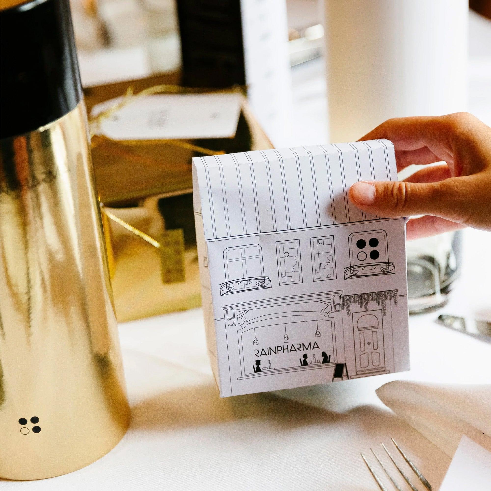 Een hand houdt een gevouwen, geïllustreerd papier vast met het opschrift "A P A R M A 2", naast een gouden fles en een witte tafelsetting. Het geheel roept een spa-achtige sfeer op die doet denken aan de RainPharma Bistro - Budget Gift set met Skin Wash Lemongrass &amp; After Oil 60 ml, waardoor het het perfecte cadeau is.