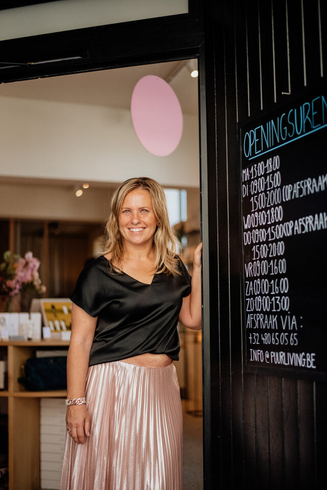 Een vrouw in een zwart topje en roze rok staat glimlachend bij de ingang van een winkel, naast een bord met de openingstijden.