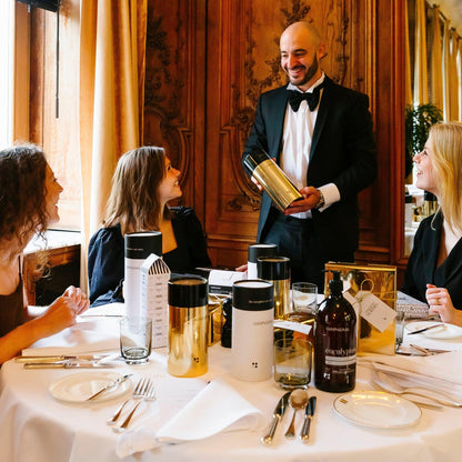 Een ober in smoking overhandigt een fles aan drie zittende vrouwen die genieten van hun maaltijd in een RainPharma Bistro, omringd door schalen en bakjes in een elegante eetkamer, wat een spa-achtig gevoel oproept. Ze waarderen de RainPharma Bistro - Budget Gift set met Skin Wash Lemongrass &amp; After Oil 60 ml.