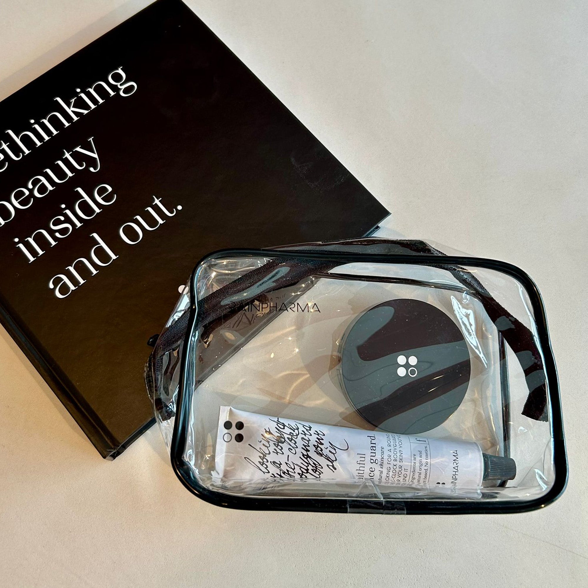 Een RainPharma Gratis toilettasje met 2 fullsize producten ligt op een tafel naast een zwart boek met de titel &quot;Rethinking beauty inside and out&quot;.