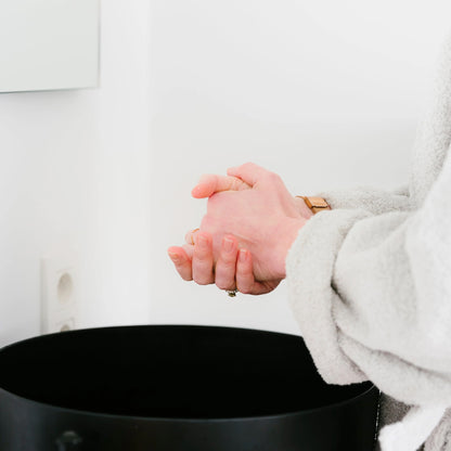 Een persoon in een lichtgrijze badjas staat bij een zwarte wastafel in een witte badkamer, klaar om RainPharma Exceptional Hand &amp; Nail Cream aan te brengen voor een zachte, gevoede huid.