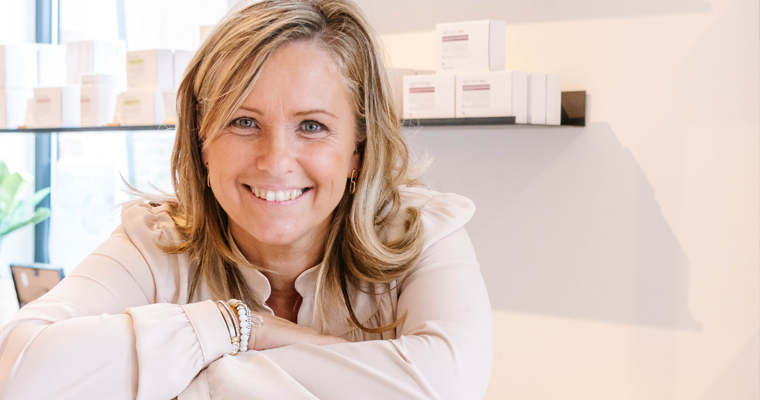 Een vrouw met schouderlang blond haar lacht naar de camera en leunt met haar armen op een toonbank in een licht kantoor of winkel met rekken vol producten achter haar.