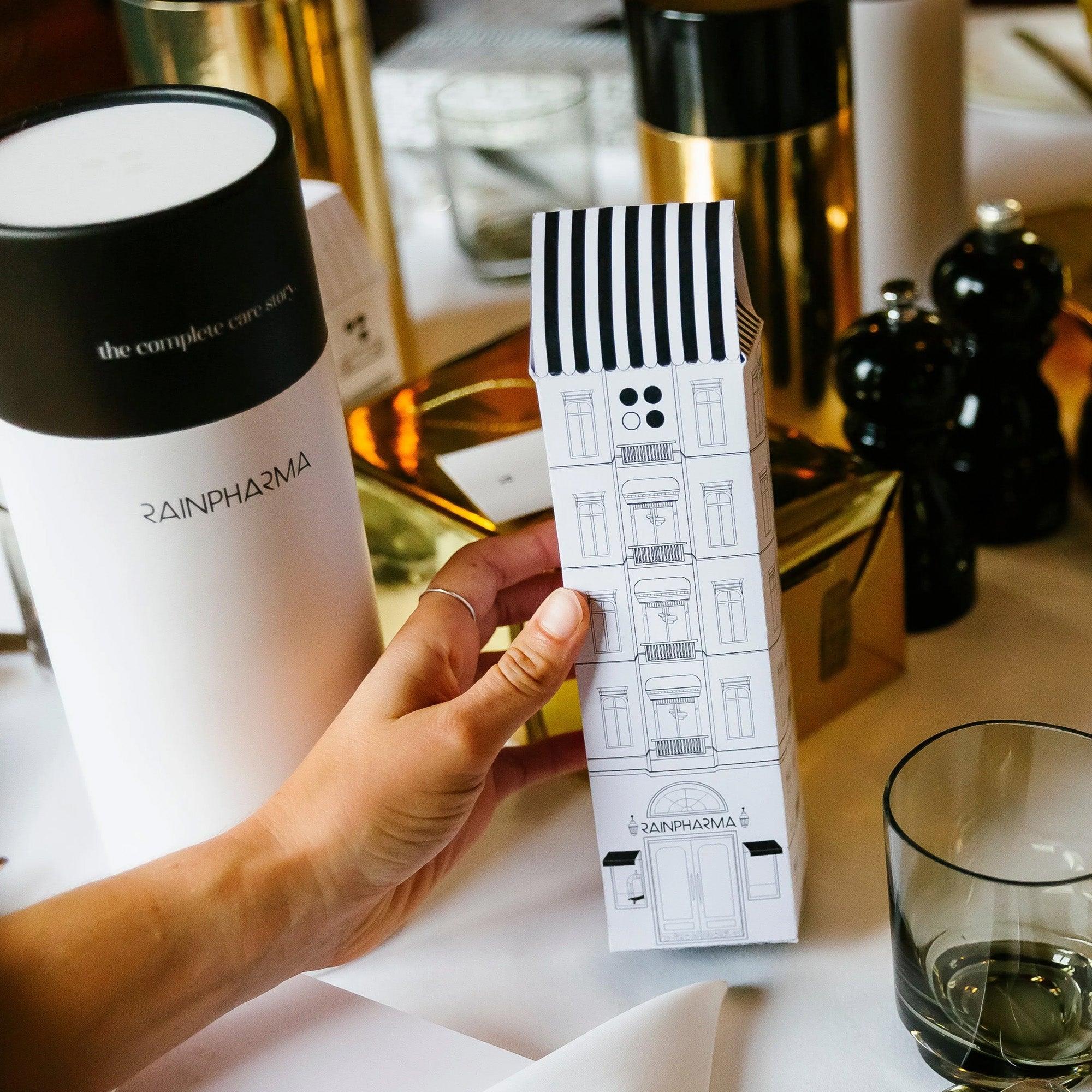Een hand houdt een RainPharma-productdoos vast, het ontwerp lijkt op een geïllustreerd gebouw, met de branding "RAINPHARMA". In de buurt liggen items zoals natuurlijke douchegel en elegant servies, wat de verfijning van de scène versterkt. De geschenkset bevat Skin Wash Lemongrass &amp; Sacred Seven Body Repair Oil 60 ml.