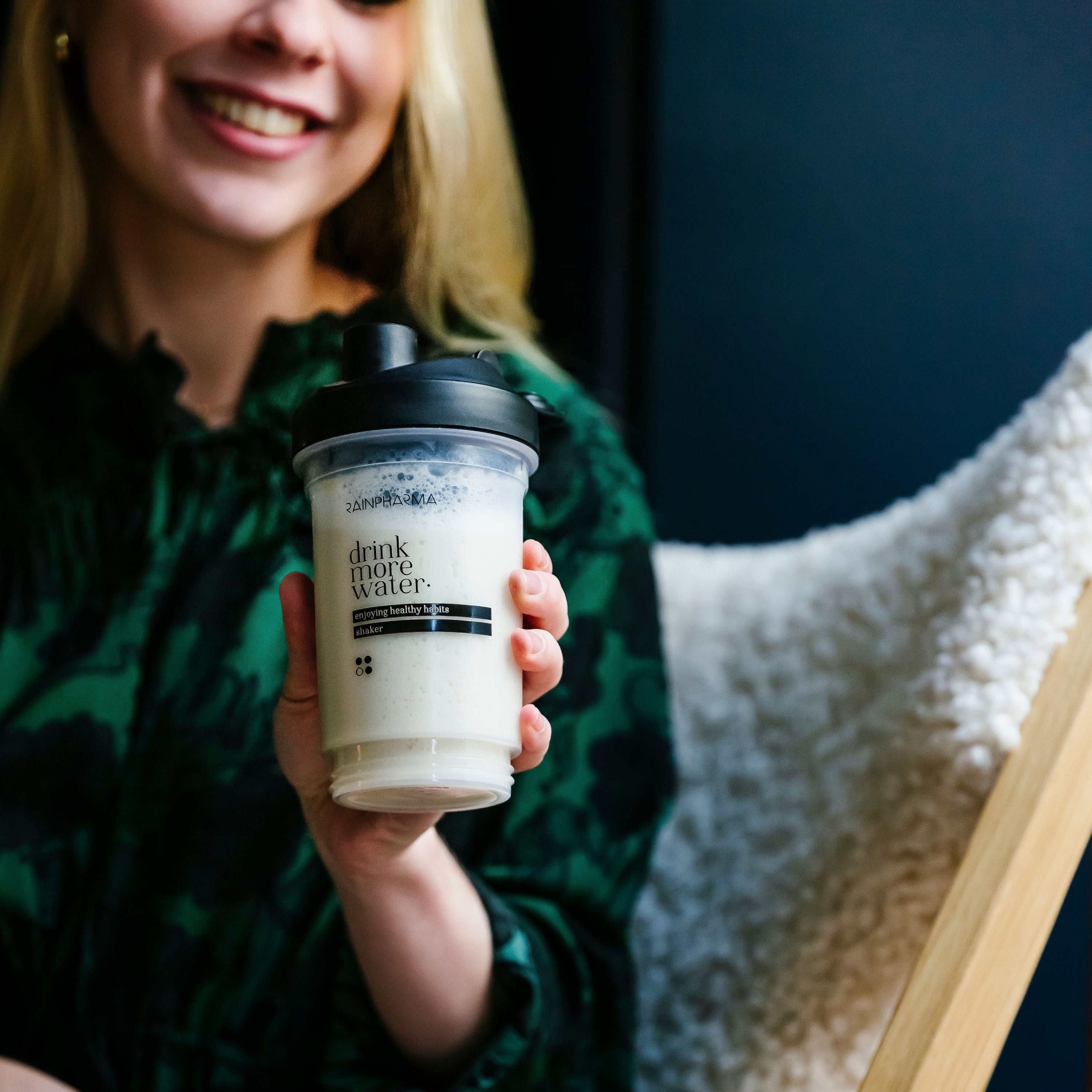 Een lachende persoon houdt een doorzichtige shaker vast met een zwarte dop waarop staat "drink meer water". Binnenin zitten de Shake Blissful Banana Zakjes van RainPharma, een lichte, eiwitrijke bananensnack gemaakt van niet-GMO-soja.