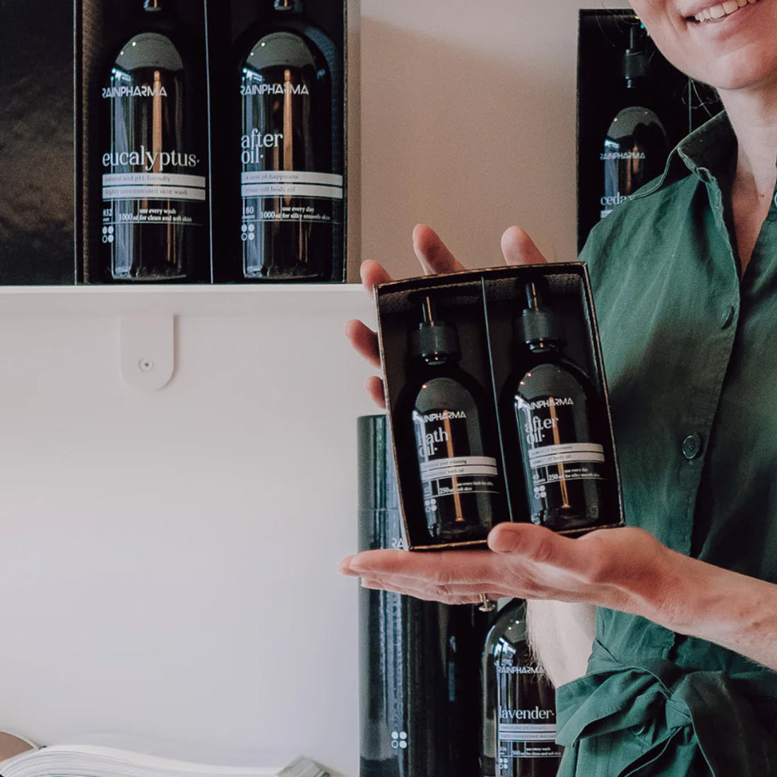 Een persoon in een groen shirt houdt een luxe RainPharma &quot;Mix &amp; Match - Stel zelf je cadeau samen&quot; geschenkdoos vast met twee donkere &quot;after oil&quot; flesjes, terwijl soortgelijke flesjes op schappen staan uitgestald - perfect als persoonlijk geschenk.