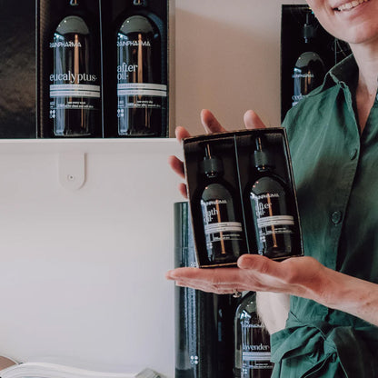 Een persoon in een groen shirt houdt een luxe RainPharma &quot;Mix &amp; Match - Stel zelf je cadeau samen&quot; geschenkdoos vast met twee donkere &quot;after oil&quot; flesjes, terwijl soortgelijke flesjes op schappen staan uitgestald - perfect als persoonlijk geschenk.