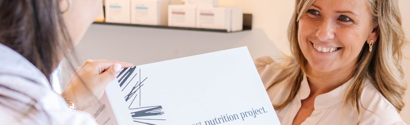 Twee vrouwen bespreken een document met de titel "voedingsproject" in een helder, modern kantoor met productdozen op een plank op de achtergrond.