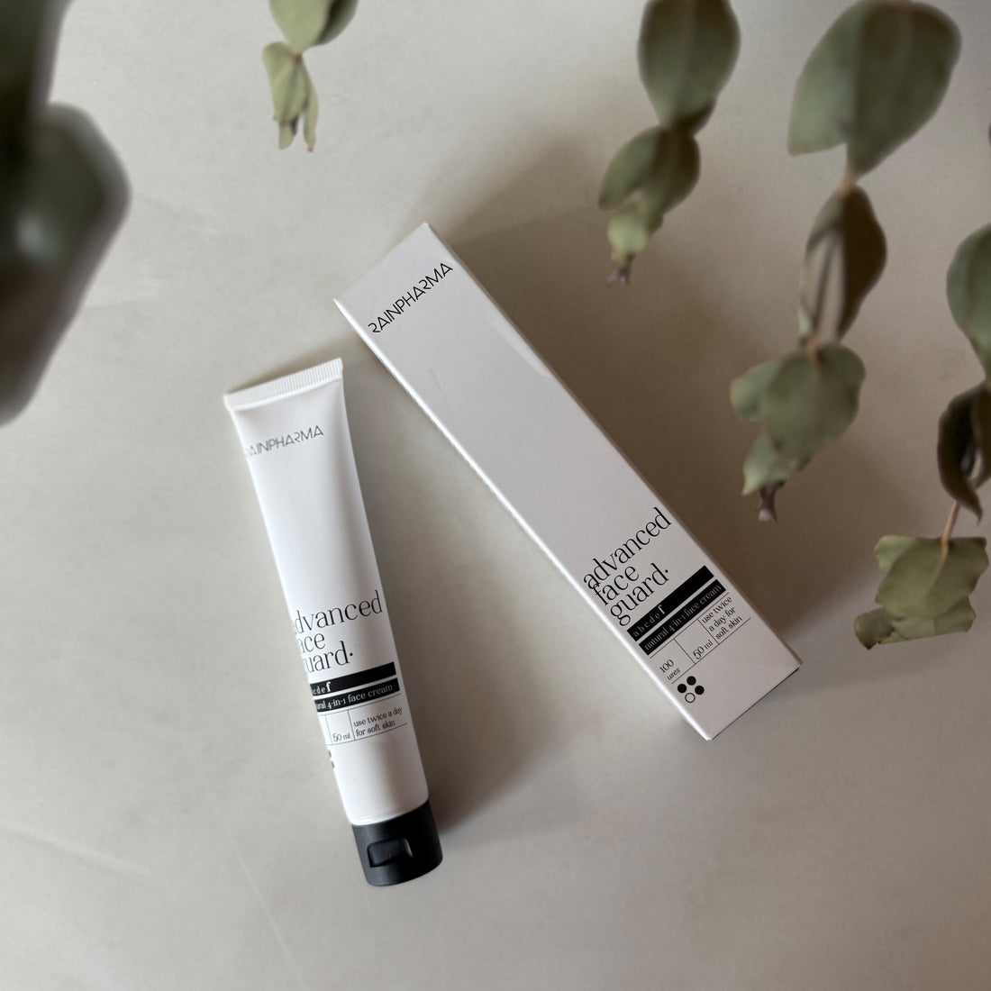 Een tube en het bijbehorende doosje met de tekst &quot;RainPharma Advanced Face Guard - NIEUW&quot; liggen op een licht oppervlak met groene bladeren in de buurt en laten de 4-in-1 gelaatscrème met rode algenextract voor geavanceerde huidverzorging zien.