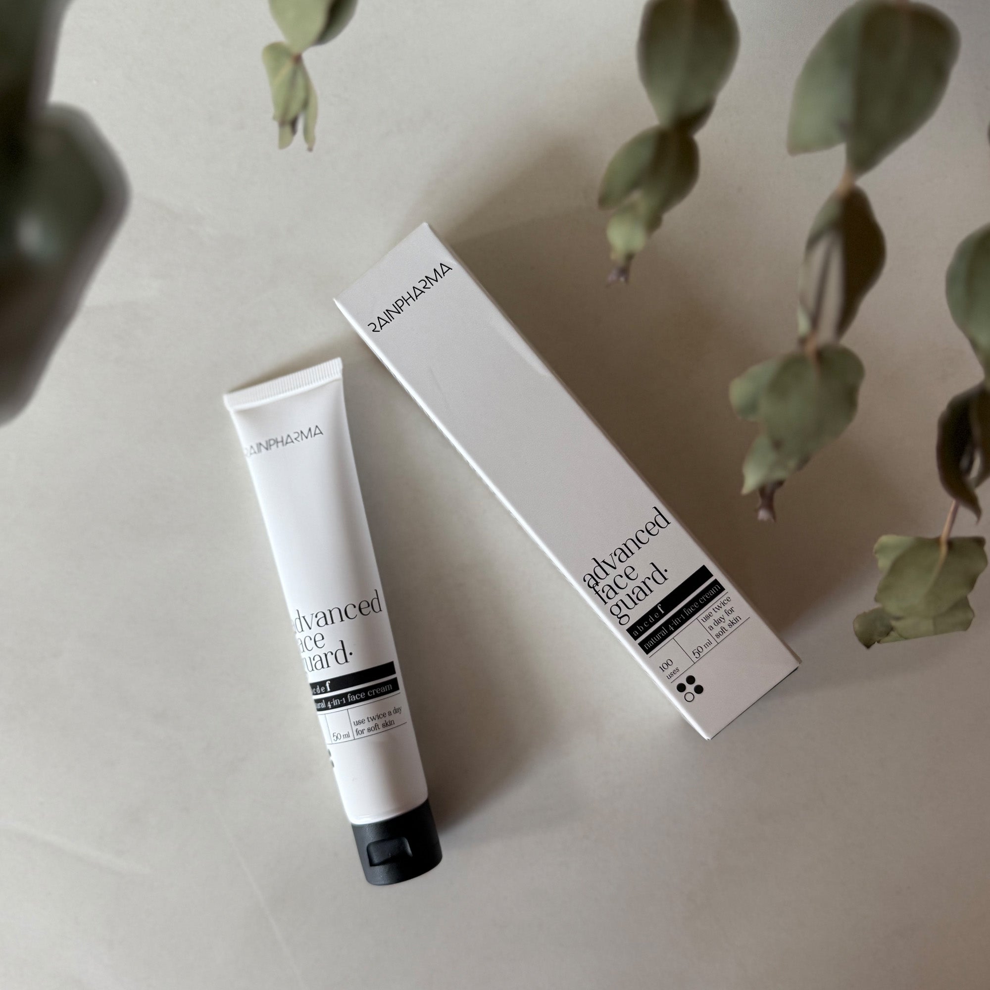 Een tube en het bijbehorende doosje met de tekst "RainPharma Advanced Face Guard - NIEUW" liggen op een licht oppervlak met groene bladeren in de buurt en laten de 4-in-1 gelaatscrème met rode algenextract voor geavanceerde huidverzorging zien.