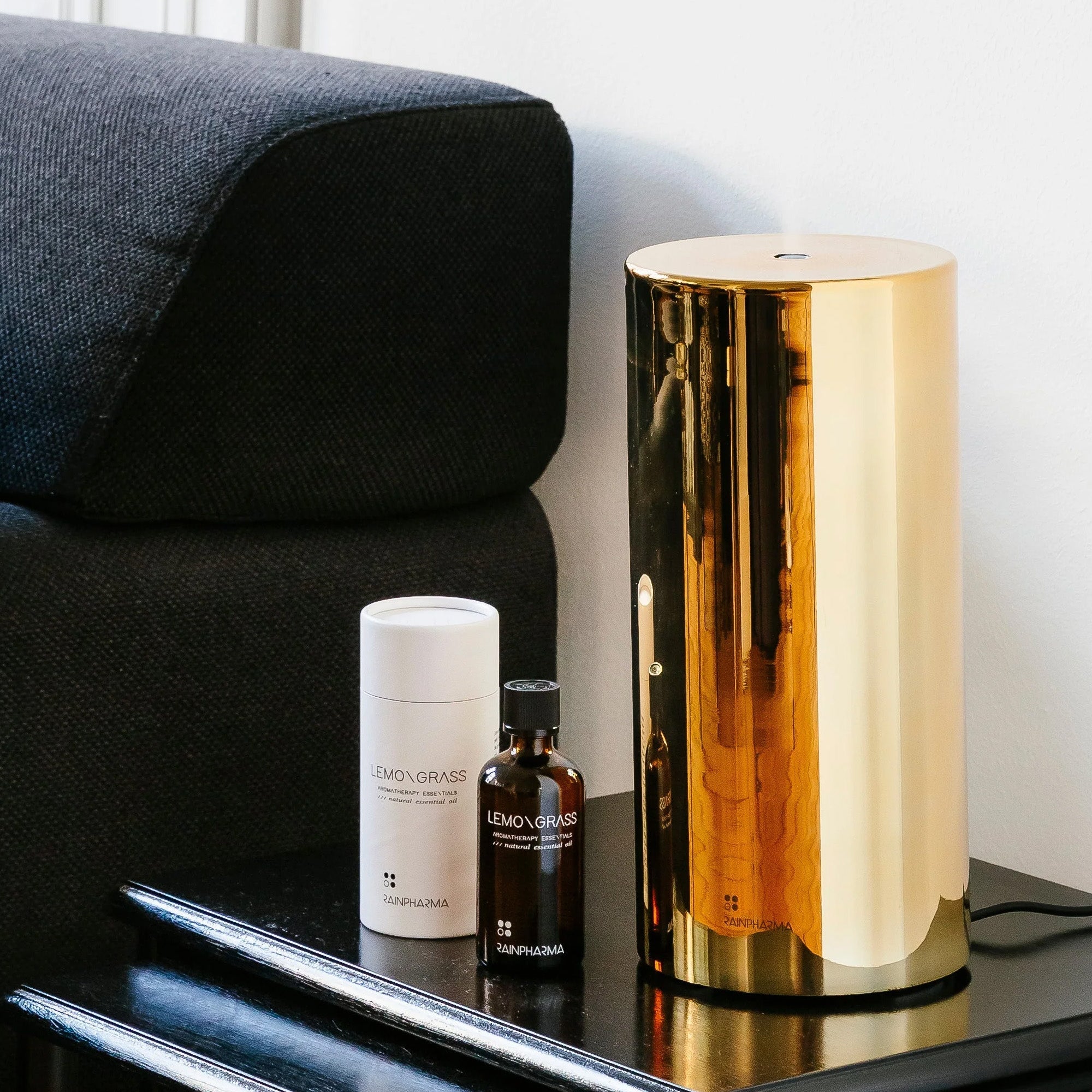 De RainPharma Aroma Diffuser Set - Aurora Base + Glass Sleeve in goud staat elegant op een zwarte tafel naast een flesje etherische olie van citroengras en de verpakking. Een donkere bank vormt een contrast met de verfijnde opstelling.