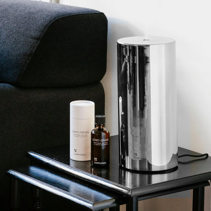 De Rainpharma Aroma Diffuser 1,3 L - Aurora Glass Sleeve staat elegant op een klein zwart tafeltje naast witte en bruine flessen en werpt sfeervol licht. Op de achtergrond is gedeeltelijk een donkere bank zichtbaar, die harmonieus opgaat in het tafereel.