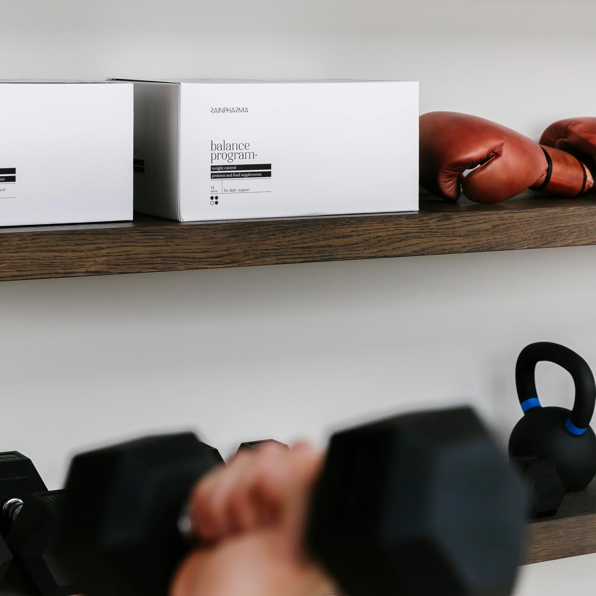 Een hand houdt een halter vast op de voorgrond; op schappen achter staan dozen met het RainPharma Balance Program (Dieet Programma), bokshandschoenen, proteïneshakes, een kettlebell en extra halters.