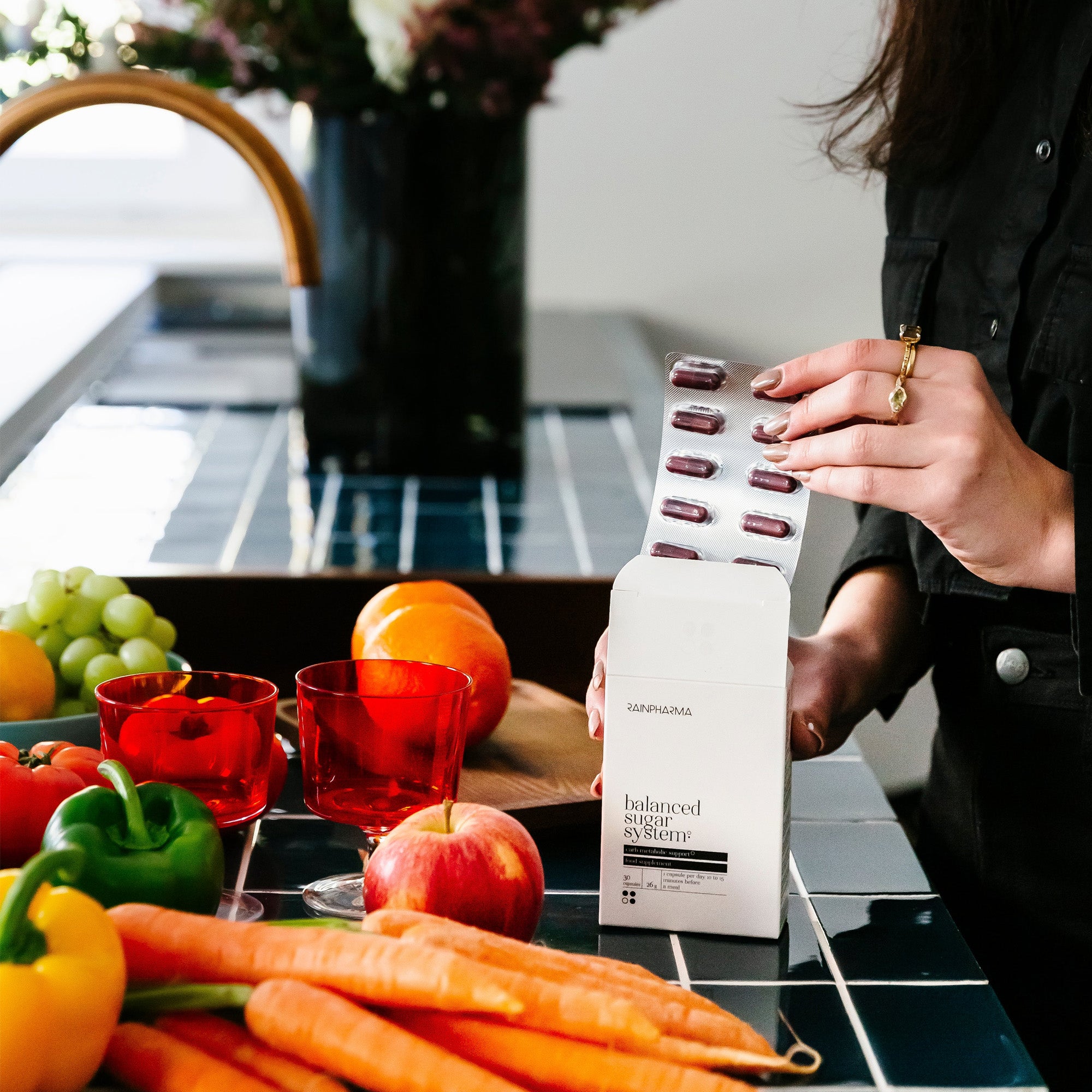 Iemand haalt een blisterverpakking capsules uit een doosje van het RainPharma Balance Program (Dieet Programma) op een aanrecht met fruit, groenten, proteïneshakes en twee rode glazen - om de focus van RainPharma op natuurlijke voeding te benadrukken.