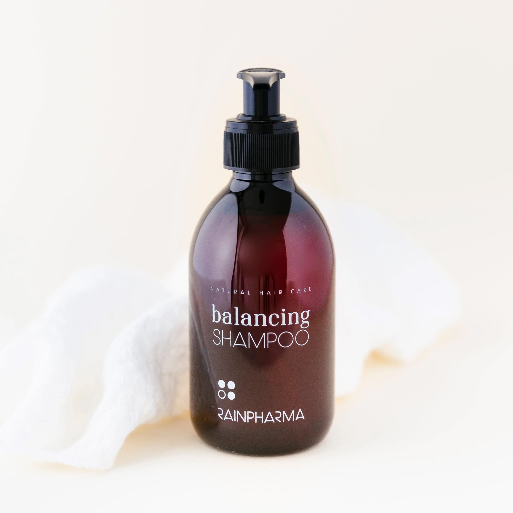 Een bruine pompfles van RainPharma Balancing Shampoo staat op een wit oppervlak met een witte doek erachter, perfect voor glanzend haar en zacht voor de gevoelige hoofdhuid.