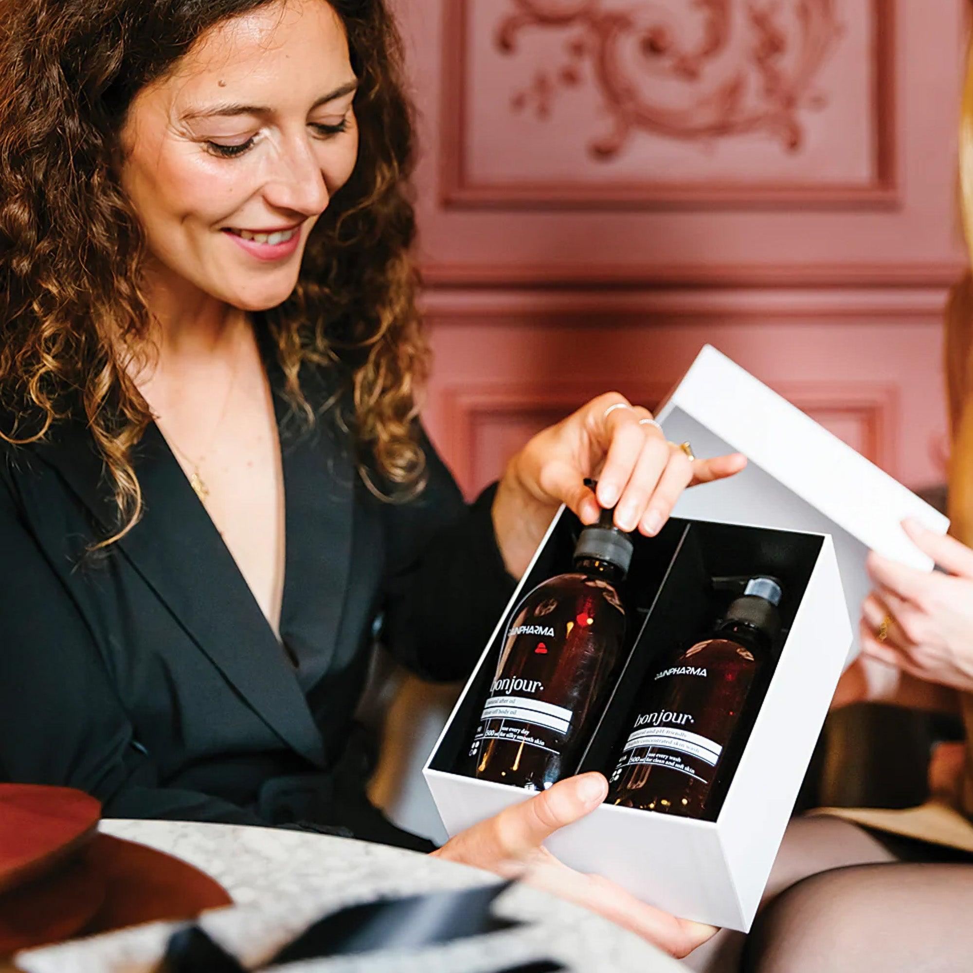 Een vrouw in het zwart glimlacht terwijl ze een witte RainPharma geschenkdoos met het Bonjour Shower Duo - Huidspoeling Bonjour en After Oil Bonjour 500ml - opent aan een tafel met een andere persoon, klaar voor een energieke start.