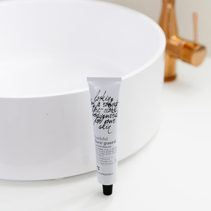 Een tube &quot;Faithful Face Guard (dag- en nachtcrème)&quot; van RainPharma met Tamanu-olie staat rechtop voor een witte, ronde wastafel met koperen kraan.