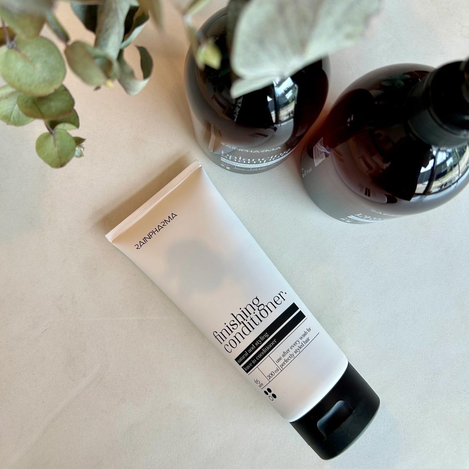 Een witte tube RainPharma Finishing Conditioner (Authentic Conditioner) rust op een licht oppervlak naast twee donkere flessen en groene bladeren, die de hydraterende eigenschappen van het product benadrukken.