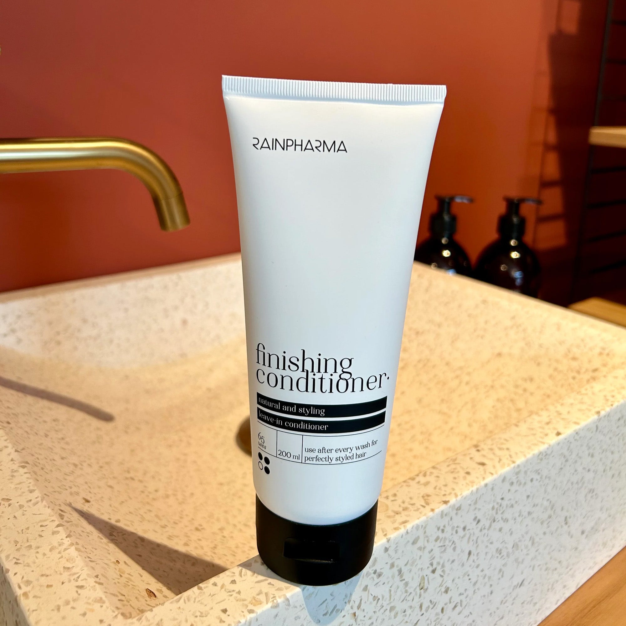 Een tube Finishing Conditioner (Authentic Conditioner) van RainPharma staat rechtop op een beige aanrecht met een gouden kraan en bruine muur en biedt een hydraterende conditionerervaring voor zijdezacht haar.