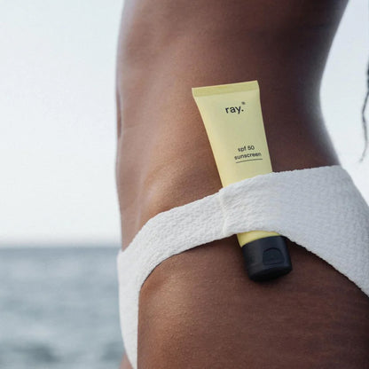 Een gele tube Ray zonnecrème SPF50 van Ray zit in de tailleband van een witte bikini met textuur, met de oceaan op de achtergrond.