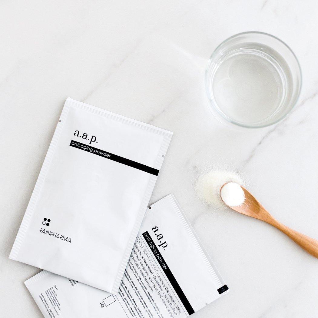 Een pakje met de naam "RainPharma Collageen Supplement - AAP (anti.aging.powder)" met chondroïtine, een glas water, een houten lepel met het poeder en een ander pakje liggen op een wit oppervlak.