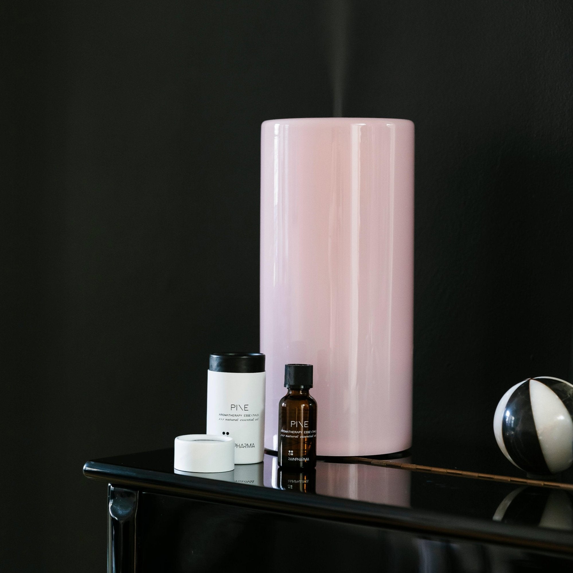 Een roze cilindervormige lamp, vergelijkbaar met de RainPharma Aroma Diffuser 1,3 L met een Aurora Glass Sleeve, staat op een glanzend zwarte tafel naast drie kleine huidverzorgingsflesjes en een decoratieve zwart-witte bol tegen een contrasterende zwarte muur.