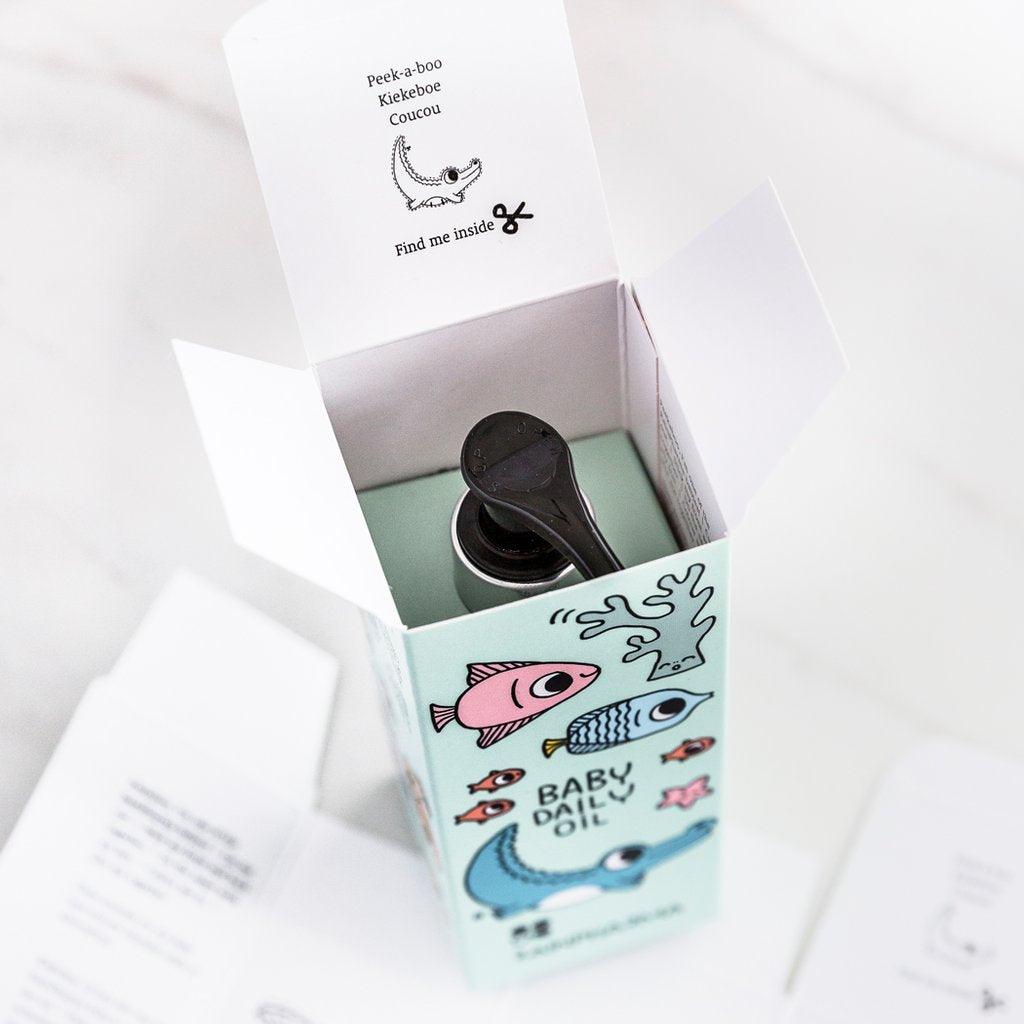 Een gedeeltelijk geopend doosje RainPharma Baby Daily Oil - 200 ml, met kleurrijke visjes en de boodschap op het deksel "Kiekeboe, vind me binnenin", naast een pictogram van een schaar - ideaal voor hypoallergene verzorging.
