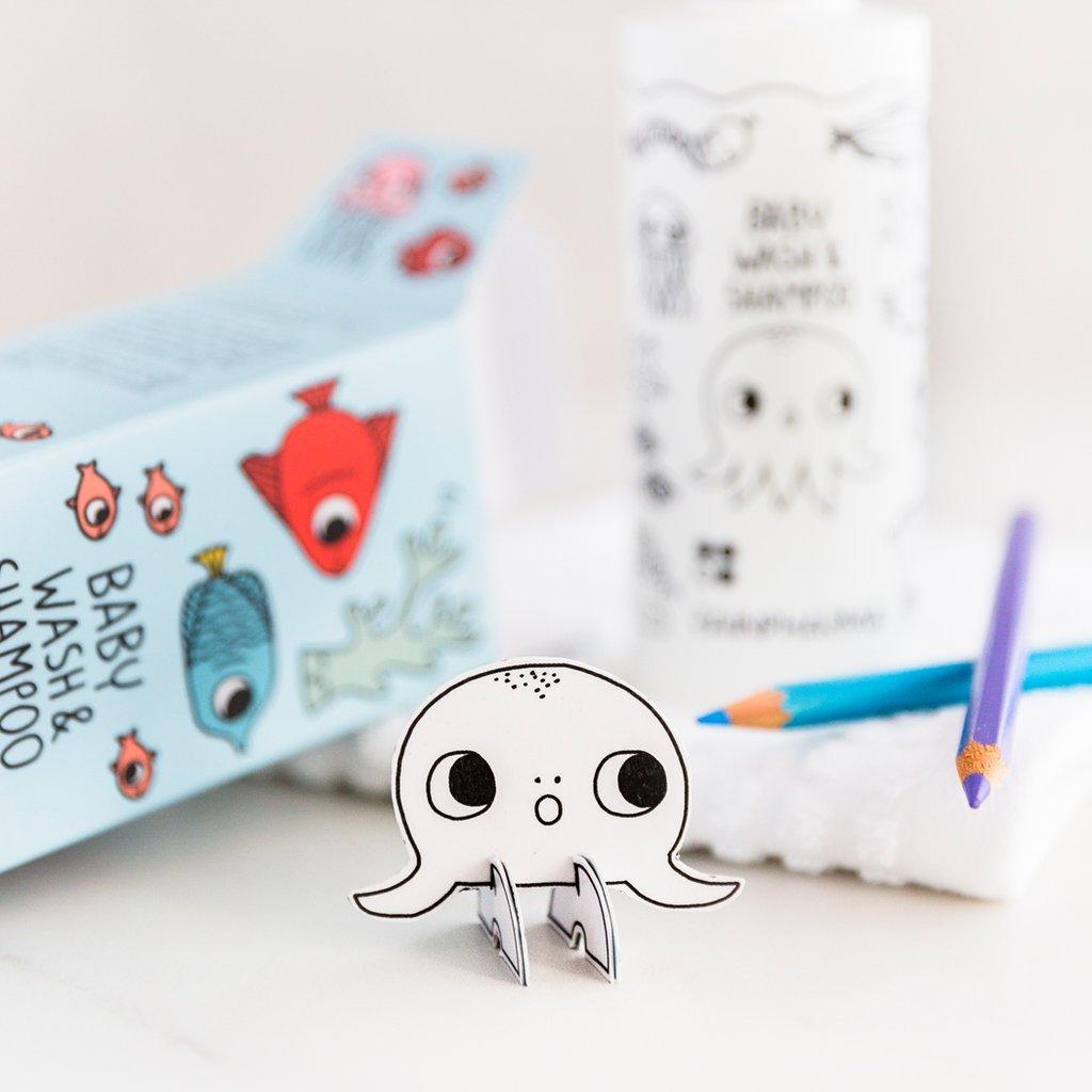 Een papieren octopus staat voor een RainPharma Baby Wash &amp; Shampoo 200 ml fles en doos, gelabeld als hypoallergene babyproducten met cartoon zeedieren, met twee kleurpotloden in de buurt.