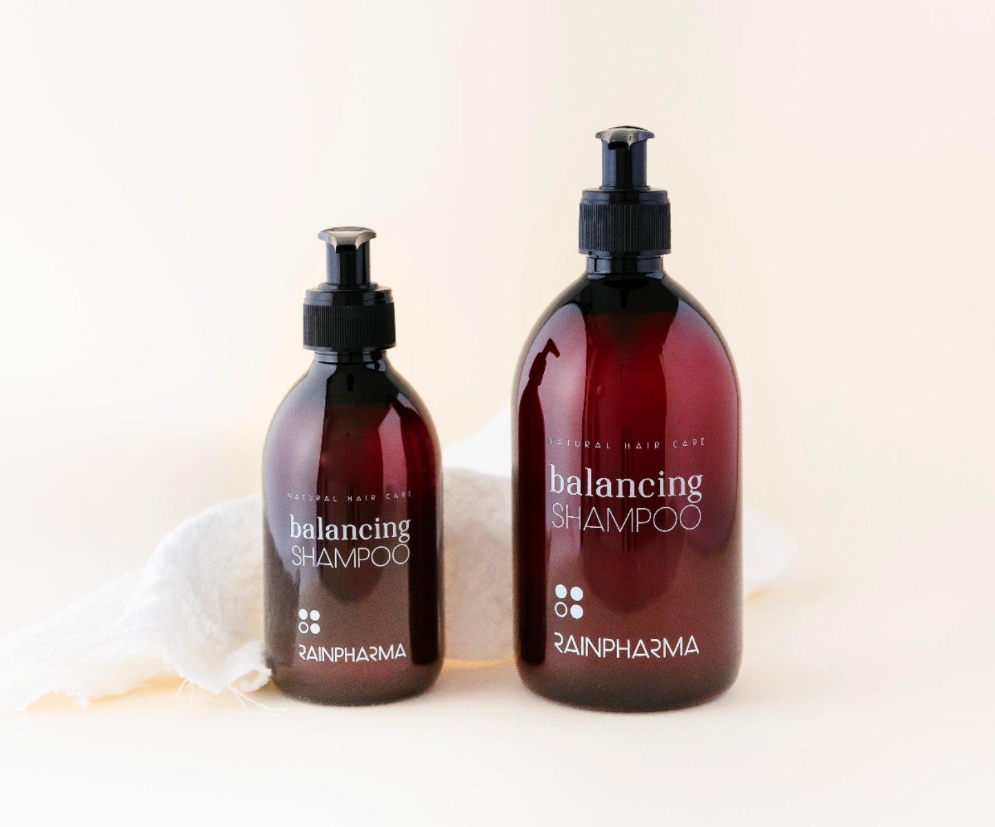 Twee amberkleurige RainPharma Balancing Shampoo-flessen met pompdispenser, perfect voor het verlichten van droge hoofdhuidproblemen, op een eenvoudige, lichte achtergrond.