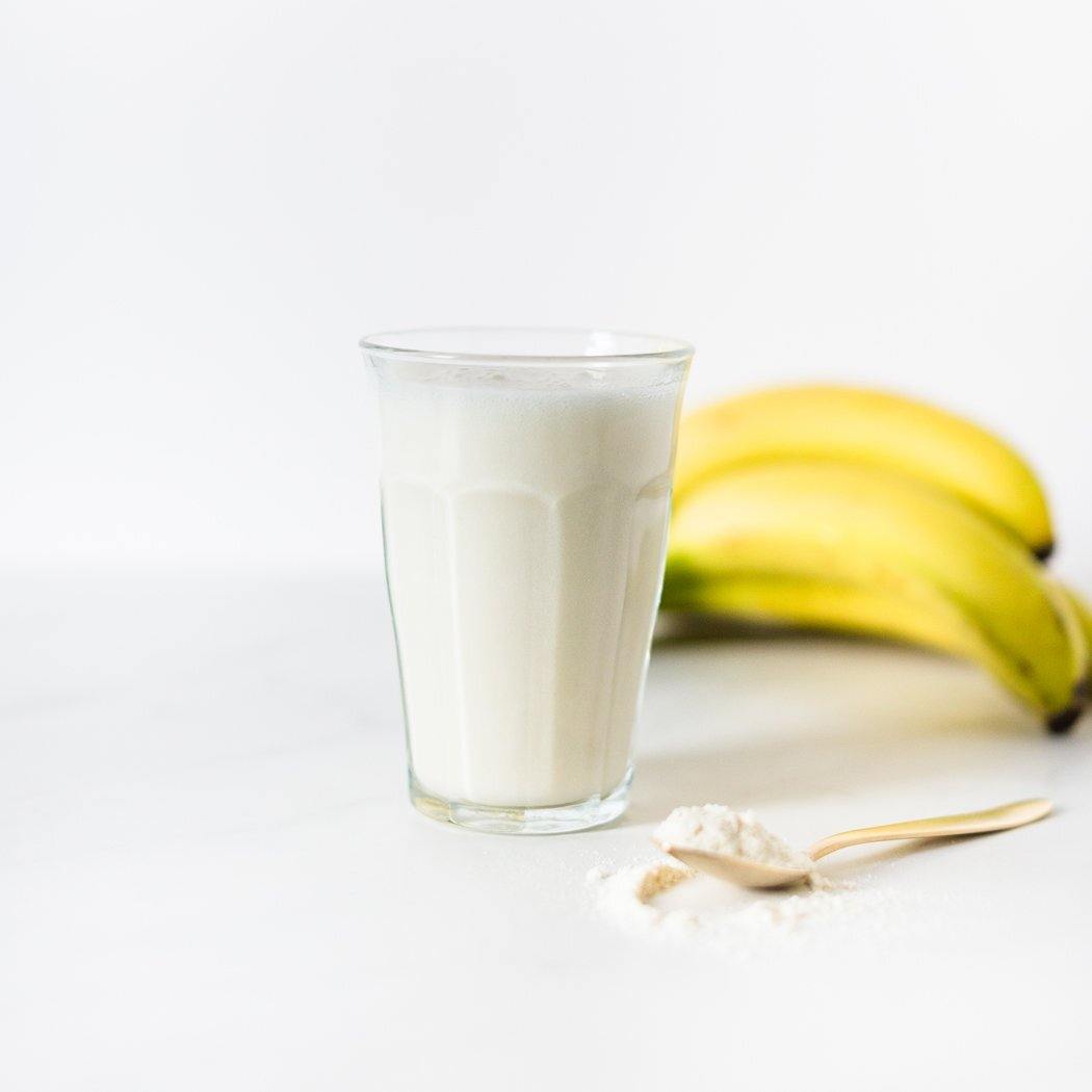 Een glas melk staat voor een tros bananen, met daarnaast een lepel poeder op een wit oppervlak. Ideaal om de Blissful Banana shake van Rainpharma te maken als eiwitrijk tussendoortje.