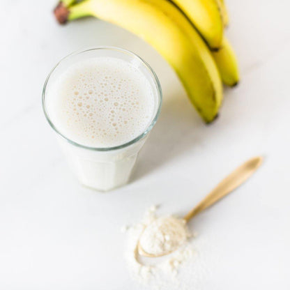 Een RainPharma Shake Blissful Banana, gemaakt met een glas melk, rijpe bananen en een lepel wit poeder, staat op een licht oppervlak - ideaal om te genieten van de bananensmaak of als eiwitrijk tussendoortje.