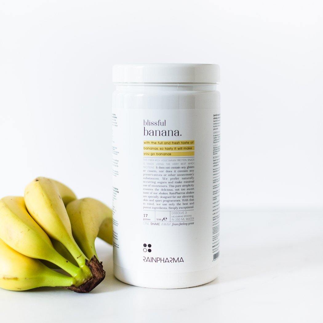 Een verpakking RainPharma Shake Blissful Banana staat naast een tros bananen op een wit oppervlak. Het is een eiwitrijke snack met een heerlijke bananensmaak.