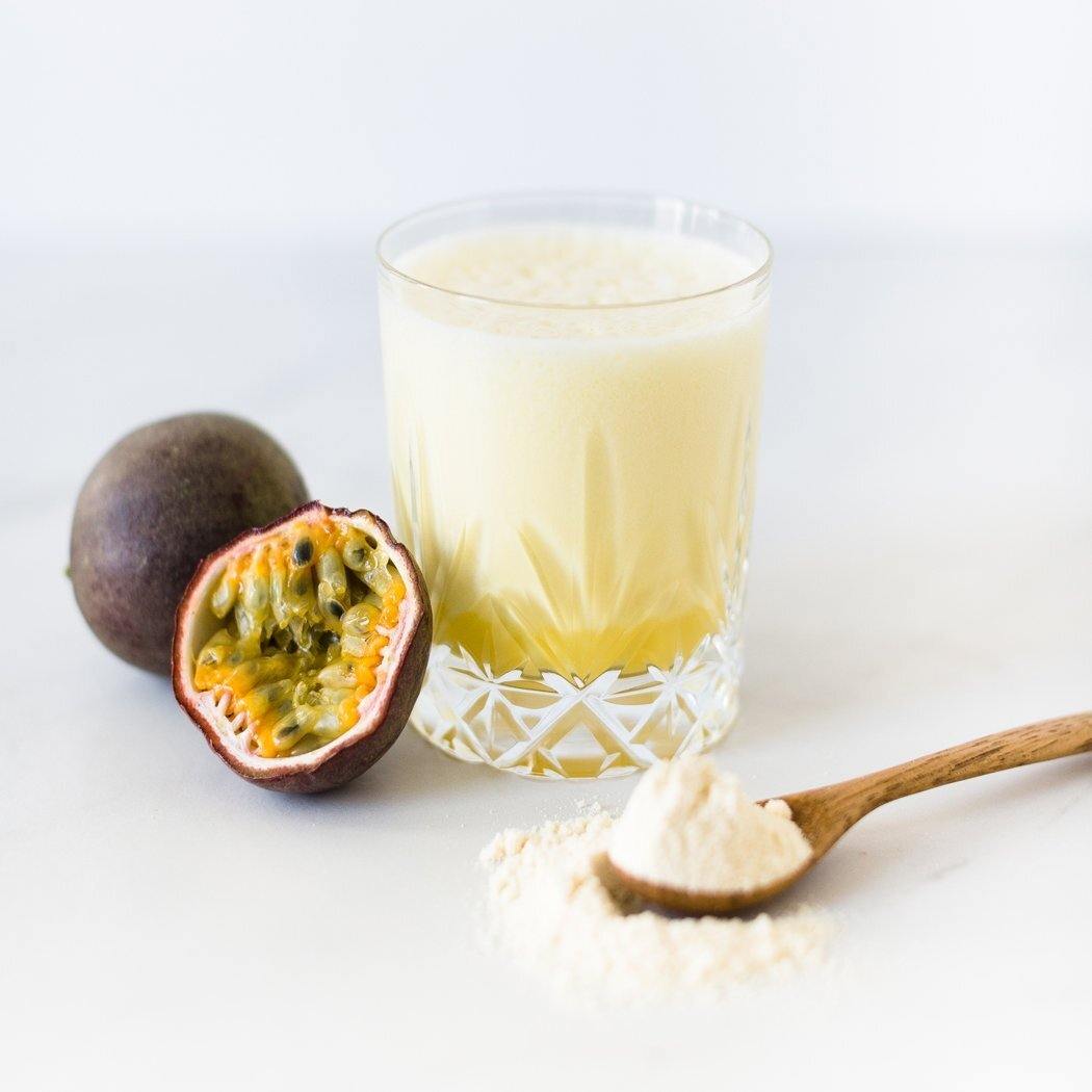 De Rainpharma Shake Brazilian Passion, boordevol proteïne en met een heerlijke passievruchten smaak, wordt elegant gepresenteerd als een gele smoothie. Het wordt vergezeld door een gehalveerde passievrucht, een hele en een lepel poeder op het maagdelijk witte oppervlak—wat het een ideaal eiwitrijk tussendoortje maakt.